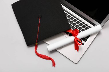 Kırmızı kurdeleli diploma, modern laptop ve beyaz arka planda mezuniyet şapkası.