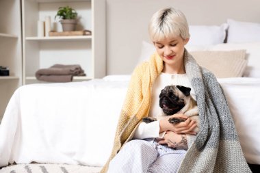 Ekose ve pug köpekli genç bir kadın yatak odasında oturuyor.