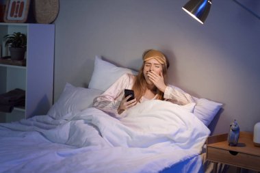 Young woman with mobile phone suffering from insomnia in bedroom at night