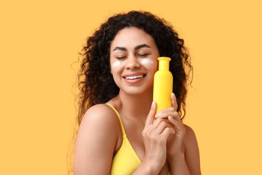 Sarı arka planda bir şişe güneş kremi olan güzel, mutlu, Afro-Amerikalı bir kadın.