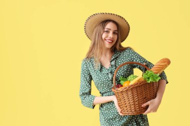 Sarı arka planda sepette piknik için yemek ve bir şişe şarapla güzel, genç bir kadın.