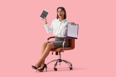 Tablet bilgisayarı ve panosu olan genç sekreter pembe arka planda ofis sandalyesinde oturuyor.