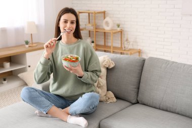 Bir kase sebze salatalı genç kadın şık bir oturma odasında kanepede oturuyor.