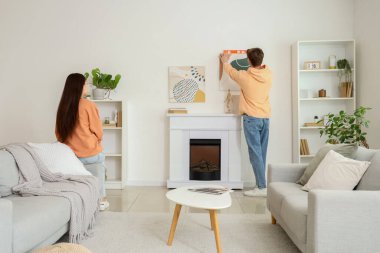 Şöminenin üstündeki duvarda asılı duran tablosu olan genç bir adam ve evde karısı, arkadan bakan.