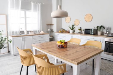 Modern mutfağın içinde beyaz tezgahlar, yemek masası, meyveler, sandalyeler ve lamba var.
