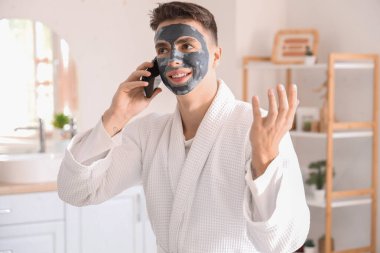 Uygulamalı kil maskeli genç adam banyoda cep telefonuyla konuşuyor.