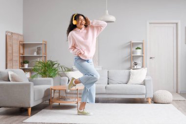 Güzel, genç, mutlu, Afro-Amerikan bir kadın kulaklıkla müzik dinliyor ve evde dans ediyor.