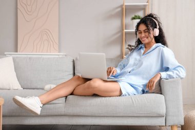 Kulaklıklı, dizüstü bilgisayarlı ve evdeki kanepede müzik dinleyen güzel, mutlu bir Afro-Amerikan kadın.