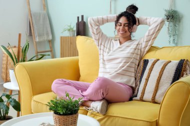 Güzel, genç, mutlu, Afro-Amerikan bir kadın kulaklıkla evdeki kanepede müzik dinliyor.