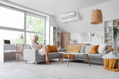 Kanepede çay içen olgun bir kadın oturma odasındaki 3 boyutlu resme bakıyor.