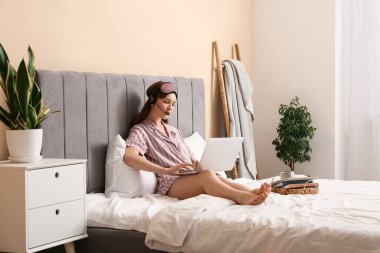Pijamalı ve kulaklıklı genç bir kadın yatak odasında dizüstü bilgisayarla çalışıyor. Uzak çalışma konsepti