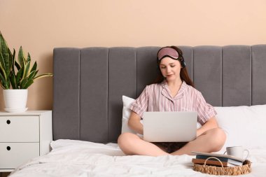 Pijamalı ve kulaklıklı genç bir kadın yatak odasında dizüstü bilgisayarla çalışıyor. Uzak çalışma konsepti