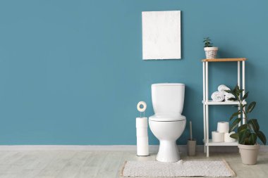 Tuvaletin içinde klozet, raf ünitesi ve mavi duvarın yanında bitkiler var.