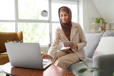 Evde tablet bilgisayar ve dizüstü bilgisayarla çalışan Müslüman iş kadını