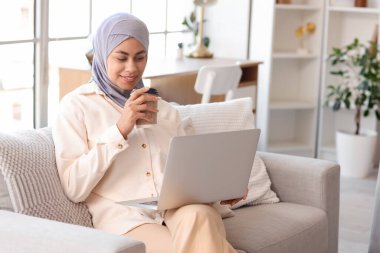 Evdeki kanepede çalışan, kahve ve dizüstü bilgisayarlı Müslüman bir iş kadını.