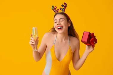 Mayo giymiş mutlu genç bir kadın ve elinde hediye kutusu ve sarı arka planda bir bardak şampanya tutan ren geyiği boynuzları. Temmuz 'da Noel