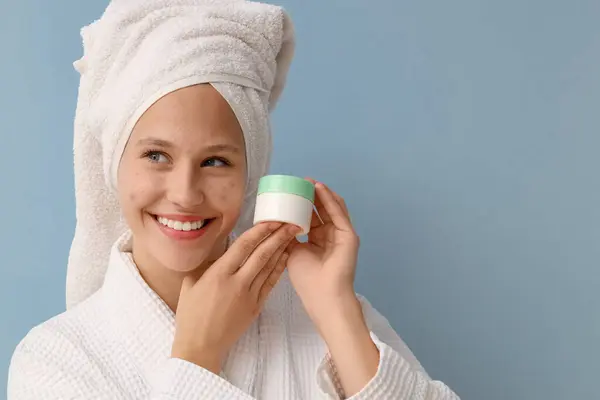stock image Beautiful young happy woman in bathrobe with acne problem and jar of cream on blue background