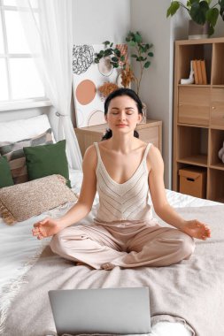 Yatak odasında dizüstü bilgisayarla meditasyon yapan güzel bir kadın.