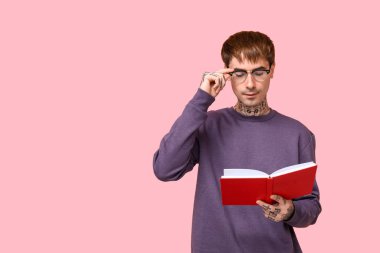 Genç dövmeli adam pembe arka planda kitap okuyor.