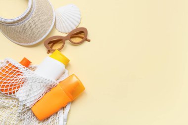 Güneş kremi şişeleri, deniz kabuğu, güneş gözlüğü ve bej arka planda şapka.