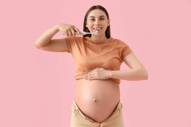 Genç hamile bir kadın pembe arka planda elektrikli diş fırçasıyla dişlerini fırçalıyor.