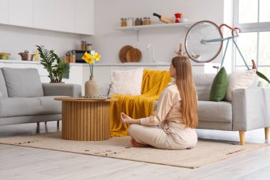 Genç bir kadın oturma odasındaki kanepenin yanında meditasyon yapıyor.
