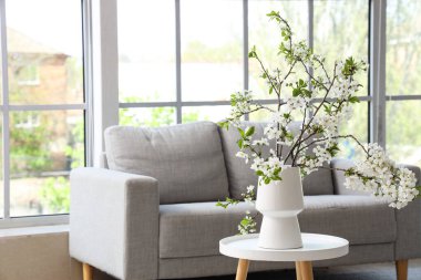 Aydınlık oturma odasındaki gri kanepenin yanındaki çekmecelerin üzerinde çiçek açan bir vazo.