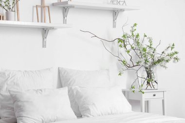 Beyaz yatak odasındaki rahat yatağın yanında, komodinin üzerinde çiçek açan dalları olan vazo.