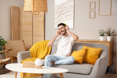 Kulaklıklı genç sakallı adam evdeki kanepede müzik dinliyor.