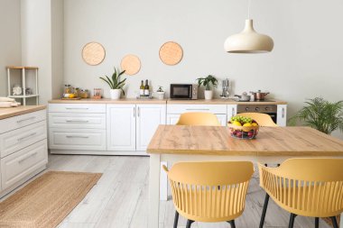 Modern mutfağın içinde beyaz tezgahlar, yemek masası, meyveler, sandalyeler ve lamba var.