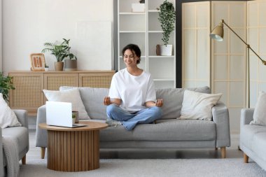 Genç Afro-Amerikalı kadın evinde dizüstü bilgisayarla kanepede meditasyon yapıyor.