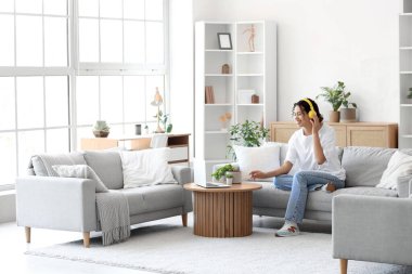 Kulaklıklı genç Afro-Amerikalı kadın evdeki kanepede ve laptopta oturuyor.