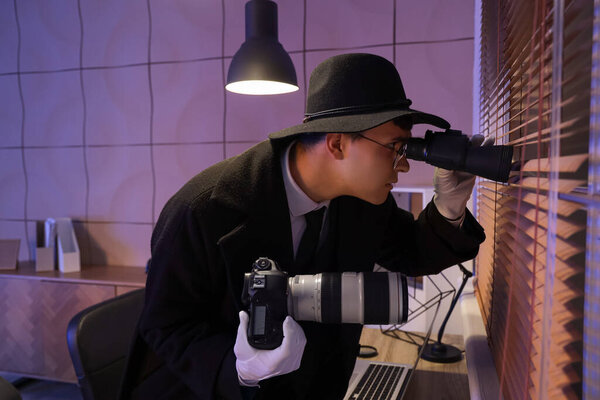 Male spy with photo camera looking through binoculars near window in office at night