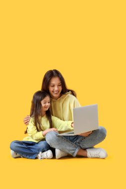 Annesi sarı arka planda dizüstü bilgisayar kullanan sevimli küçük bir kız.