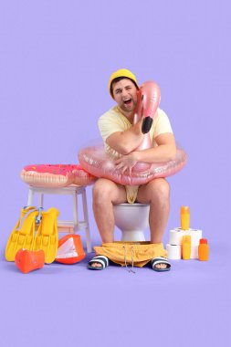 Plaj aksesuarlı bir erkek, klozetin üzerinde leylak arka planda oturuyor.