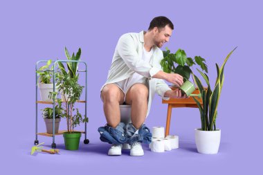 Genç adam klozete oturmuş leylak arka planda.