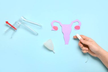 Kağıt uterusu olan kadın eli, jinekolojik spekulum, tampon ve mavi arka planda regl bardağı.