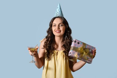 Doğum günü şapkalı genç bayan, açık mavi arka planda hediye ve kokteyl.