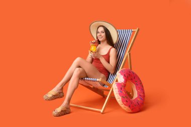 Şık kırmızı mayo giymiş, bir bardak kokteyl ve turuncu arka planda şezlongda oturan şişme yüzüklü güzel bir kadın.