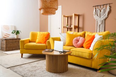 Oturma odasının içinde sarı kanepe, koltuk ve dizüstü bilgisayar ahşap masada.