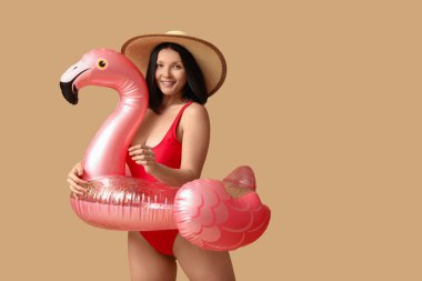 Havalı kırmızı mayo giyen, kahverengi arka planda flamingo şeklinde şişirilebilir yüzük takan güzel bir kadın.