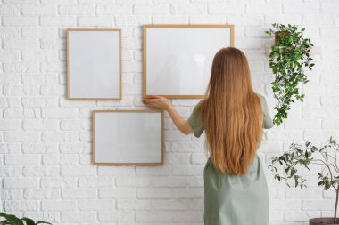 Genç bir kadın oturma odasındaki beyaz tuğla duvara boş çerçeveler asıyor.