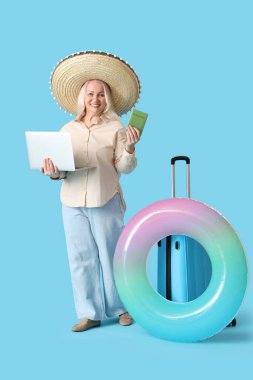 Laptop, pasaport ve yüzme yüzüğüyle şapkalı olgun bir kadın mavi arka planda yaz tatiline hazır.