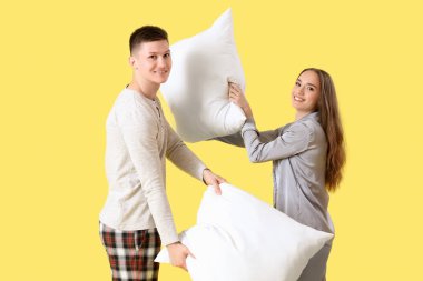 Sarı arka planda pijamalı genç bir çift yastıklarla kavga ediyor.
