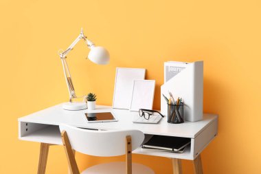 Tablet bilgisayarı, masa lambası ve turuncu duvarın yanındaki not defterleriyle rahat bir iş yeri.