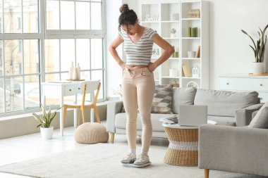 Dar pantolonlu genç Afro-Amerikalı kadın ağırlığını evde tartıyla ölçüyor. Diyet kavramı