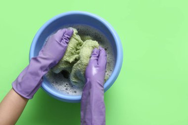 Lastik eldivenli bir kadın yeşil arka planda plastik bir leğende çamaşır yıkıyor.