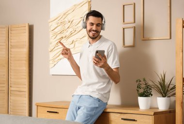 Kulaklıklı, cep telefonlu genç sakallı adam evde müzik dinliyor.