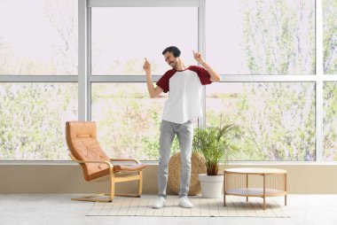 Kulaklıklı genç sakallı adam evin penceresinin yanında dans ediyor.