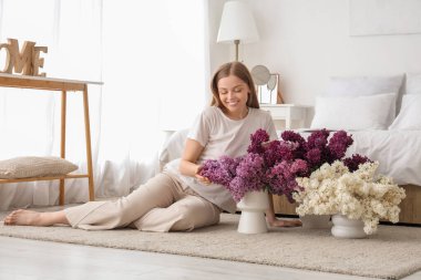 Genç bir kadın yatak odasında vazolarla yerde oturuyor.